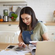 indoor shot of attractive young female doing calculations, managing home budget, sitting at kitchen table and smiling. pretty girl paying domestic bills online, using laptop computer and calculator