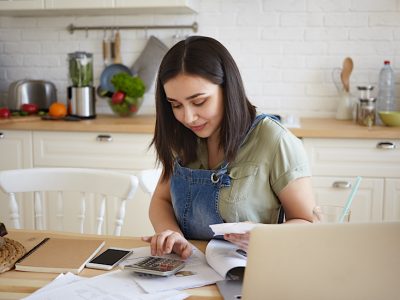 indoor shot of attractive young female doing calculations, managing home budget, sitting at kitchen table and smiling. pretty girl paying domestic bills online, using laptop computer and calculator