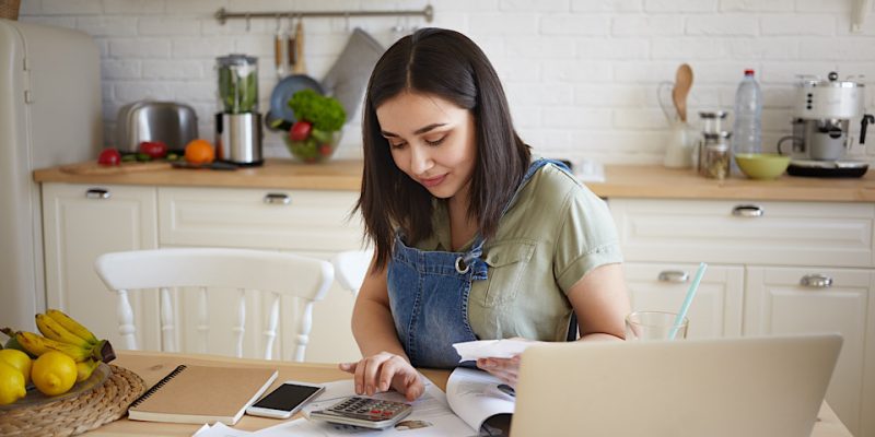 indoor shot of attractive young female doing calculations, managing home budget, sitting at kitchen table and smiling. pretty girl paying domestic bills online, using laptop computer and calculator